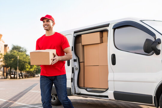 Cheerful young delivery man in red uniform carrying package cardboard box from his white van, delivering parcel to customer