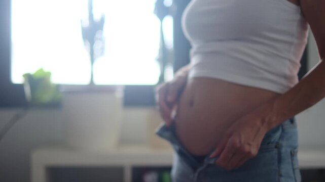 14 weeks pregnant woman struggles to button jeans due to growing belly. Embracing weight gain and self-care, promoting healthy skin and well-being at home