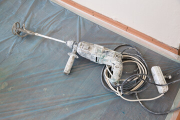Large hammer drill with mixer bit, power cable and three-way socket. A blue plastic cleaning tarpaulin is laid on the floor.