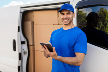 Happy delivery driver man standing beside his van full of parcel boxes, holding digital tablet,...