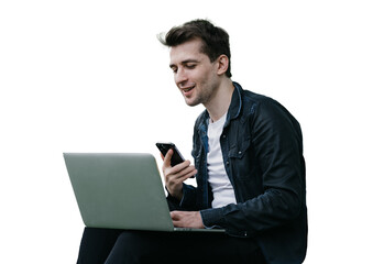 Handsome brunette cheerful male sitting on grass against transparent background with laptop making video call holds phone smiling having break remote working. man talking with friend via internet.