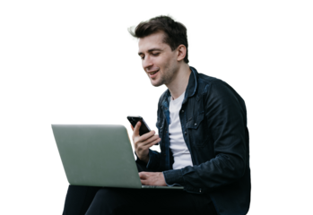 Handsome brunette cheerful male sitting on grass against transparent background with laptop making video call holds phone smiling having break remote working. man talking with friend via internet.