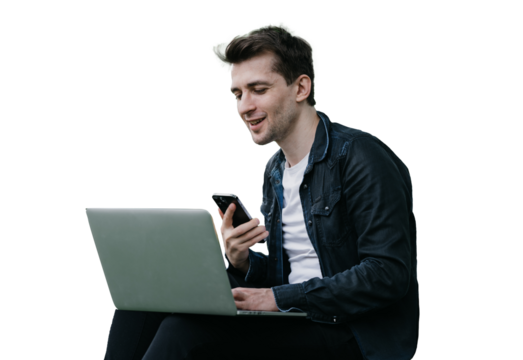 Handsome brunette cheerful male sitting on grass against transparent background with laptop making video call holds phone smiling having break remote working. man talking with friend via internet.