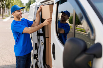 Young delivery man taking parcel off from van full of cardboard boxes and packages or loading parcels into truck