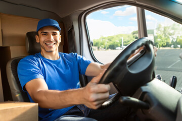 Happy young European male courier in blue uniform sitting on driver's seat of van, ready to deliver parcels, smiling at camera