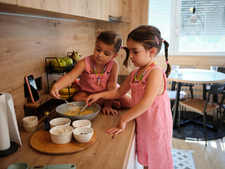 Two young girls are preparing dough for baking on the kitchen counter. Various baking ingredients...