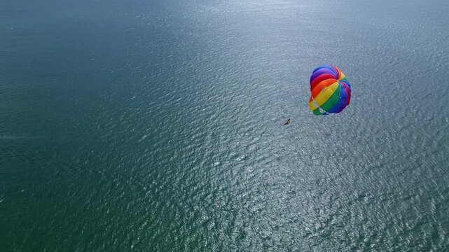 Drone video of a colorful parasail soaring over the open ocean, towed by a speedboat below. The aerial view captures the thrill and freedom of parasailing against a vast, serene seascape