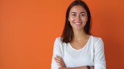 A smiling woman in a white t-shirt confidently crosses her arms against a vibrant orange backdrop, exuding positivity and approachability, her relaxed demeanor enhancing the portrait.
