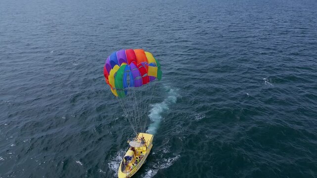 Drone video of a colorful parasail being towed by a speedboat over the ocean. The scene captures the excitement of parasailing with stunning views of the coastline and open sea
