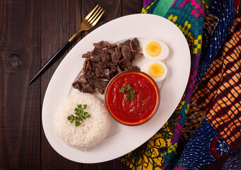 Overhead view of West African beef, white rice, boiled egg and shito sauce with an African linen napkin on a dark wooden surface; copy space