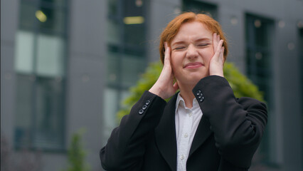 Close up stressed overworked fatigued young Caucasian redhead nervous businesswoman female woman girl elegant suit hands holding head outdoors city urban street. Job bad news work stress health issues