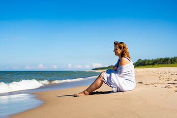 Summer holidays. Beautiful young woman in blue swimsuit covered with white scarf sitting on sand by sea on beach on sunny day. Side view.