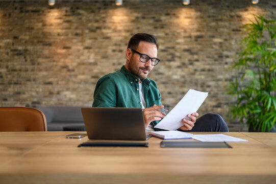 Focused male accountant in eyeglasses reading documents while working over laptop at desk in office