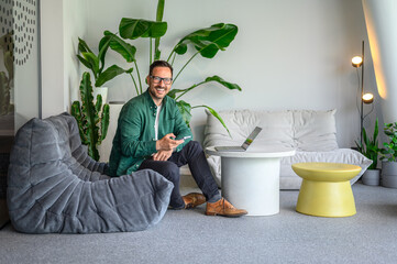 Portrait of successful male entrepreneur with smart phone and laptop sitting on sofa in modern...