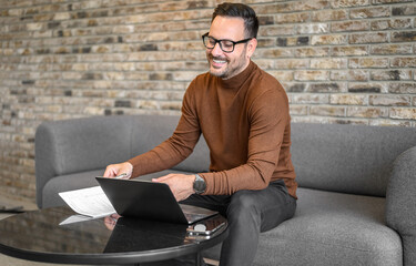 Smiling male freelancer in glasses doing online research over computer while sitting on sofa in office