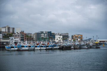 Seogwipo-si, Jeju-do, South Korea - April 25, 2022: A group of fishing boats are moored on the sea against buildings and dark clouds in the sky at Seogwipo Port