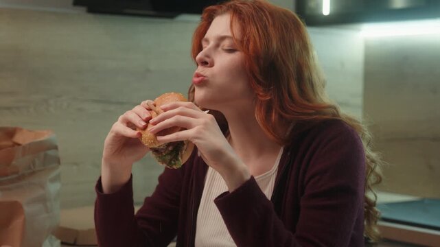 Young Caucasian woman female girl customer client eating junk fast food bite chewing burger hamburger cheeseburger eat pleasure diet cheat meal temptation indoors. Unhealthy snack overeating calories