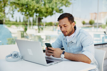 Young man working remotely at an outdoor cafe, focused on his smartphone and laptop, enjoying a sunny day