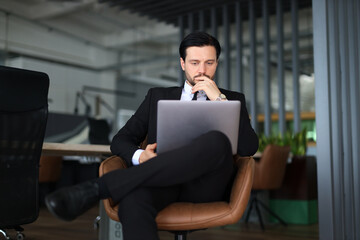 A man in a suit is sitting in a chair with a laptop in front of him. He is focused on his work, possibly typing or browsing the internet. Concept of professionalism and productivity