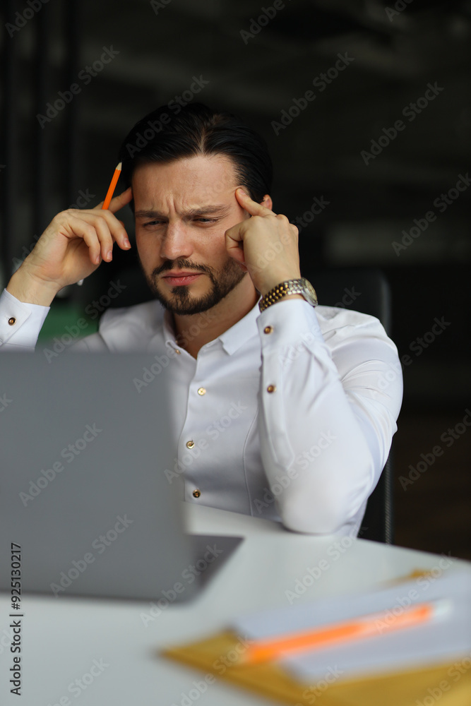 Poster a man is sitting at a desk with a laptop and a stack of papers. he is looking at the laptop and he i
