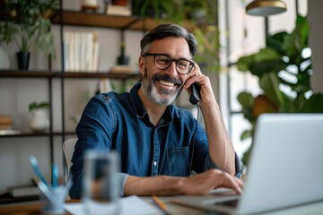 Obraz premium Smiling businessman wearing glasses talking on phone, sitting at desk with laptop, friendly manager consulting customer by phone, happy man chatting with friends distracted from work