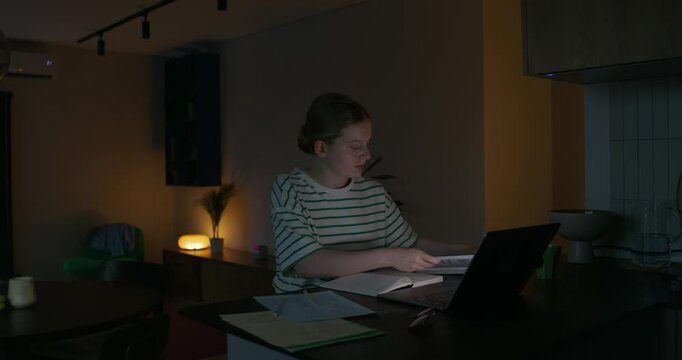 A woman is reading a book and writing in a notebook while sitting at a table in front of an open laptop. Dark room, home interior