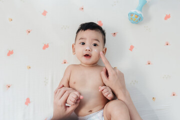 Happy Asian Baby with a Comfortable Smile, Mother Finger Gently Applying Skin Care Lotion on His...