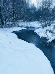 Snow-covered banks along the blue surface of the stream. Winter landscape of freezing water....