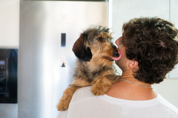 A small dachshund kisses and licks his master's face with affection and both show an attitude of good owner-pet relationship. They are inside the kitchen of a house