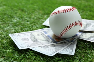 Baseball ball and dollar banknotes on green grass outdoors, closeup