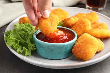 Woman dipping nugget into chili sauce at grey table, closeup