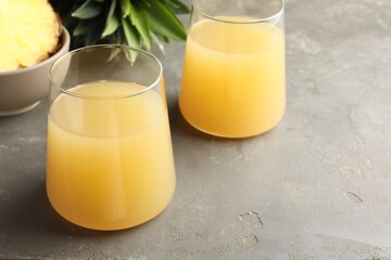 Tasty pineapple juice in glasses and fruits on grey textured table, closeup