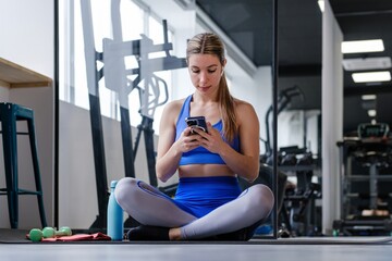A woman in a blue sports outfit attentively uses her smartphone, likely tracking her fitness progress or planning her training routine