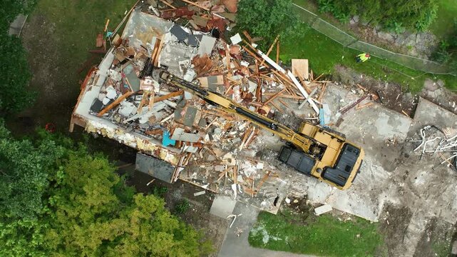 Heavy machinery demolition excavator scrapes as claw spins and pulls together large pile of refuse from broken walls and beams, drone top down orbit