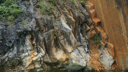 An image of a rocky cliff with a mix of gray and reddish hues, surrounded by greenery.