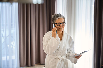 Happy adult lady in bathrobe with documents talking on the smartphone in hotel room