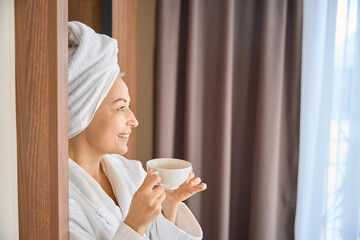 Smiling adult woman wearing bathrobe drinking tea standing in hotel room