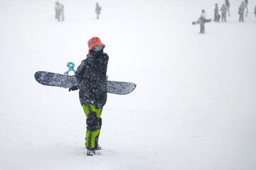 Snowboarding in any weather. A woman in a hat and sunglasses with a snowboard behind her back going up the ski slope in heavy snowfall.