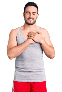 Young handsome man wearing swimwear and sleeveless t-shirt smiling with hands on chest with closed eyes and grateful gesture on face. health concept.