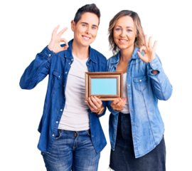 Couple of women holding empty frame smiling positive doing ok sign with hand and fingers. successful expression.