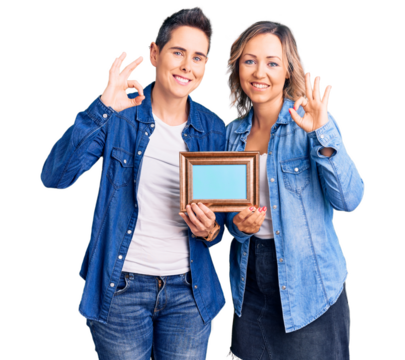 Couple of women holding empty frame smiling positive doing ok sign with hand and fingers. successful expression.
