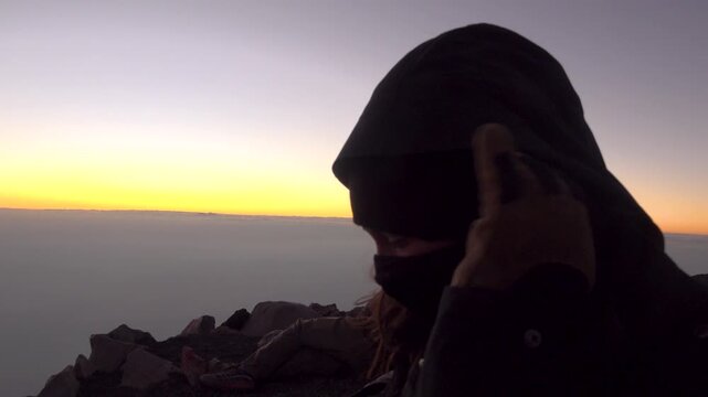 A Woman puts on her neck gaiter and looks at Fuego volcano erupting. Above the clouds at sunset in Guatemala