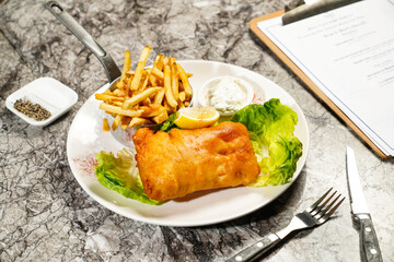 Fried cod fish and chips with tartar sauce with lemon, fork and knife served in plate isolated on marble background side view of hong kong food