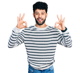 Naklejka premium Young arab man with beard wearing casual striped sweater looking surprised and shocked doing ok approval symbol with fingers. crazy expression
