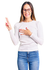 Beautiful brunette young woman wearing casual white sweater and glasses smiling swearing with hand on chest and fingers up, making a loyalty promise oath