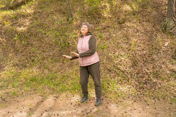 A woman dressed in a pink vest and dark pants is playfully gesturing outdoors, surrounded by trees and greenery.