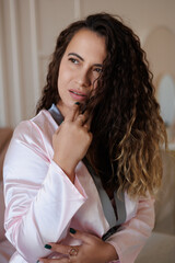 A beautiful curly-haired girl in a pink silk pajamas lies on the bed, relaxed. A middle-aged woman is getting ready for bed in the morning, smiling. Girl has fun on her bed at home, jumping