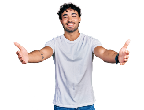 Hispanic young man with beard wearing casual white t shirt looking at the camera smiling with open arms for hug. cheerful expression embracing happiness.