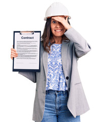Young beautiful brunette woman wearing architect hardhat holding contract stressed and frustrated with hand on head, surprised and angry face