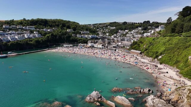 Looe Beach with coastal buildings, parked cars, many beachgoers and green cliffs on a sunny day